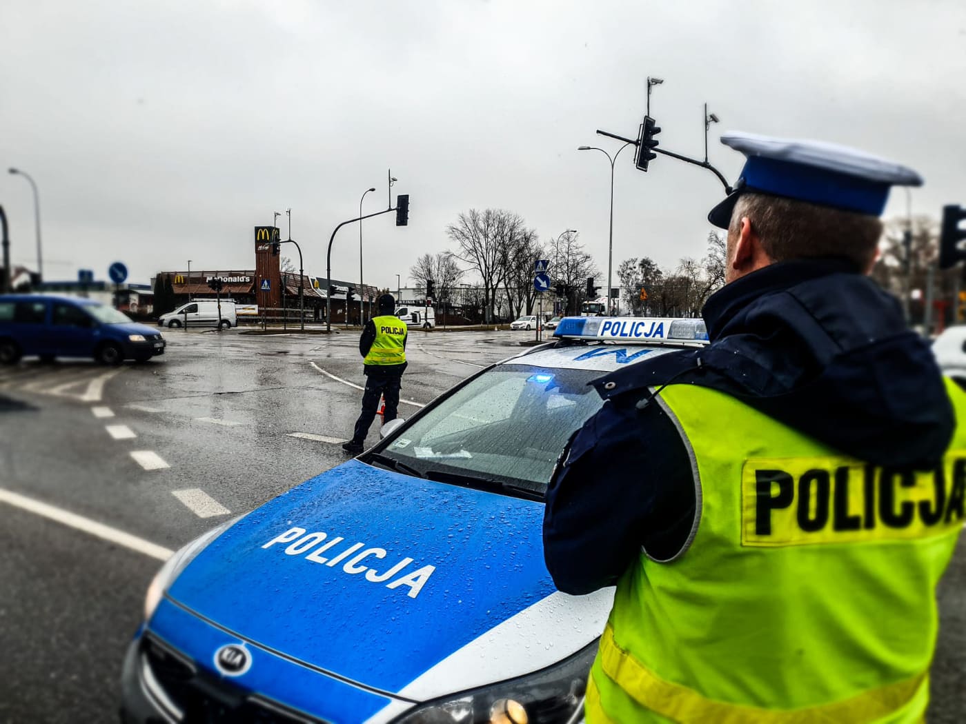 Na skrzyżowaniu stoi oznakowany policyjny samochód. Przed samochodem, tyłem do patrzącego na zdjęcie stoi funkcjonariusz w odblaskowej kamizelce z czarnym napisem: Policja