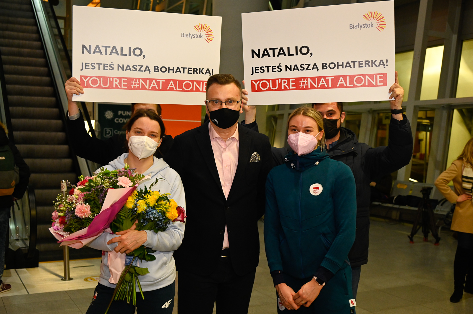 Pomiędzy dwiema kobietami w sportowych strojach stoi mężczyzna w ciemnej marynarce i jasno różowej koszuli . Za nimi stoją dwie osoby, które trzymają tabliczki z napisami: Natalio, jesteś naszą bohaterką. Pod spodem na czerwonym pasku biały napis : You're #Nat Alone