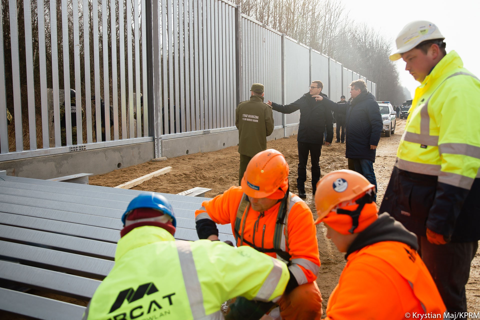 Na pierwszym planie pracownicy w odblaskowych zielono-pomarańczowych ubraniach wierzchnich i kaskach składają stalową zaporę. W tle widać fragment już postawionej stalowej zapory, obok, której stoi trzech mężczyzn, którzy rozmawiają między sobą i pokazują palcem na ogrodzenie