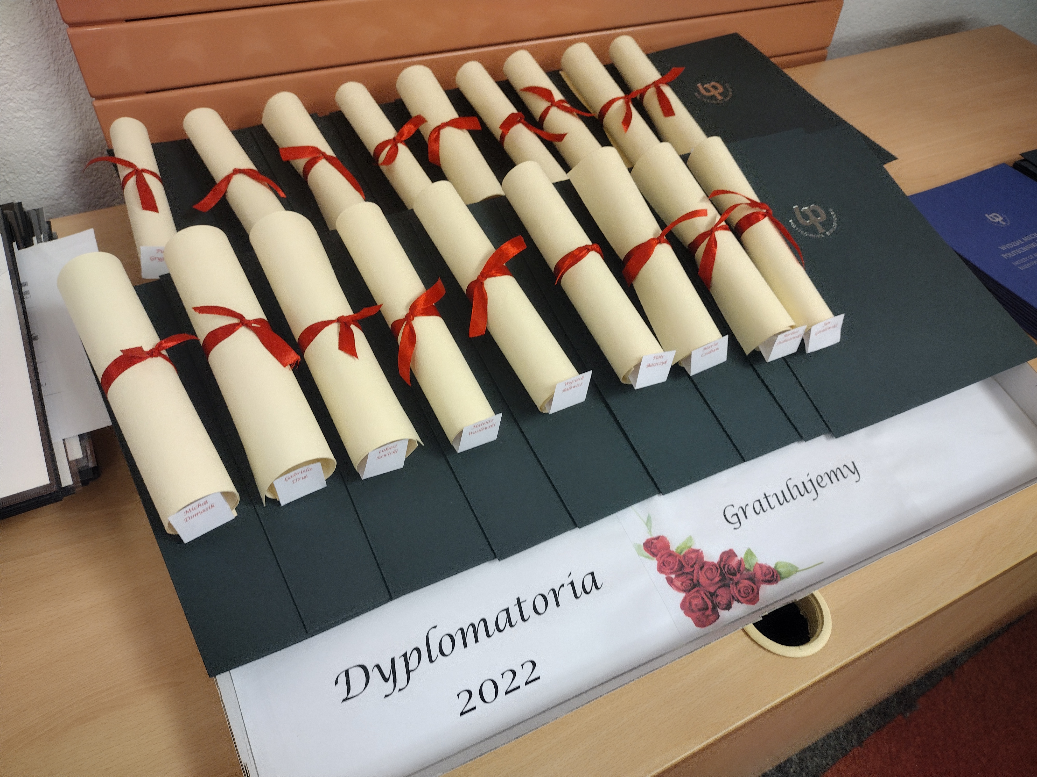 Na stole leżą w kremowym kolorze, zwinięte w rulon, przewiązane czerwoną wstążką listy gratulacyjne. Listy położone są na zielonych teczkach, w których znajdują się dyplomy ukończenia studiów. Pod teczkami znajduje się biała kartka, na której znajduje się czarny napis: Dyplomatoria 2022. Gratulujemy