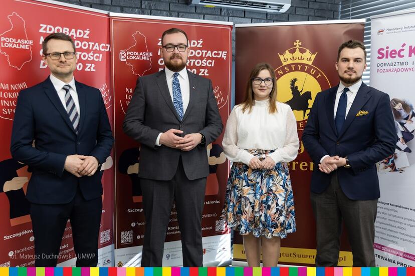 Na tle czerwonych, brązowych i białych banerów stoją cztery osoby. Od lewej strony dwóch mężczyzn w ciemnych garniturach, kobieta w kwiecistej spódnicy i białej koszuli oraz mężczyzna w ciemnym garniturze
