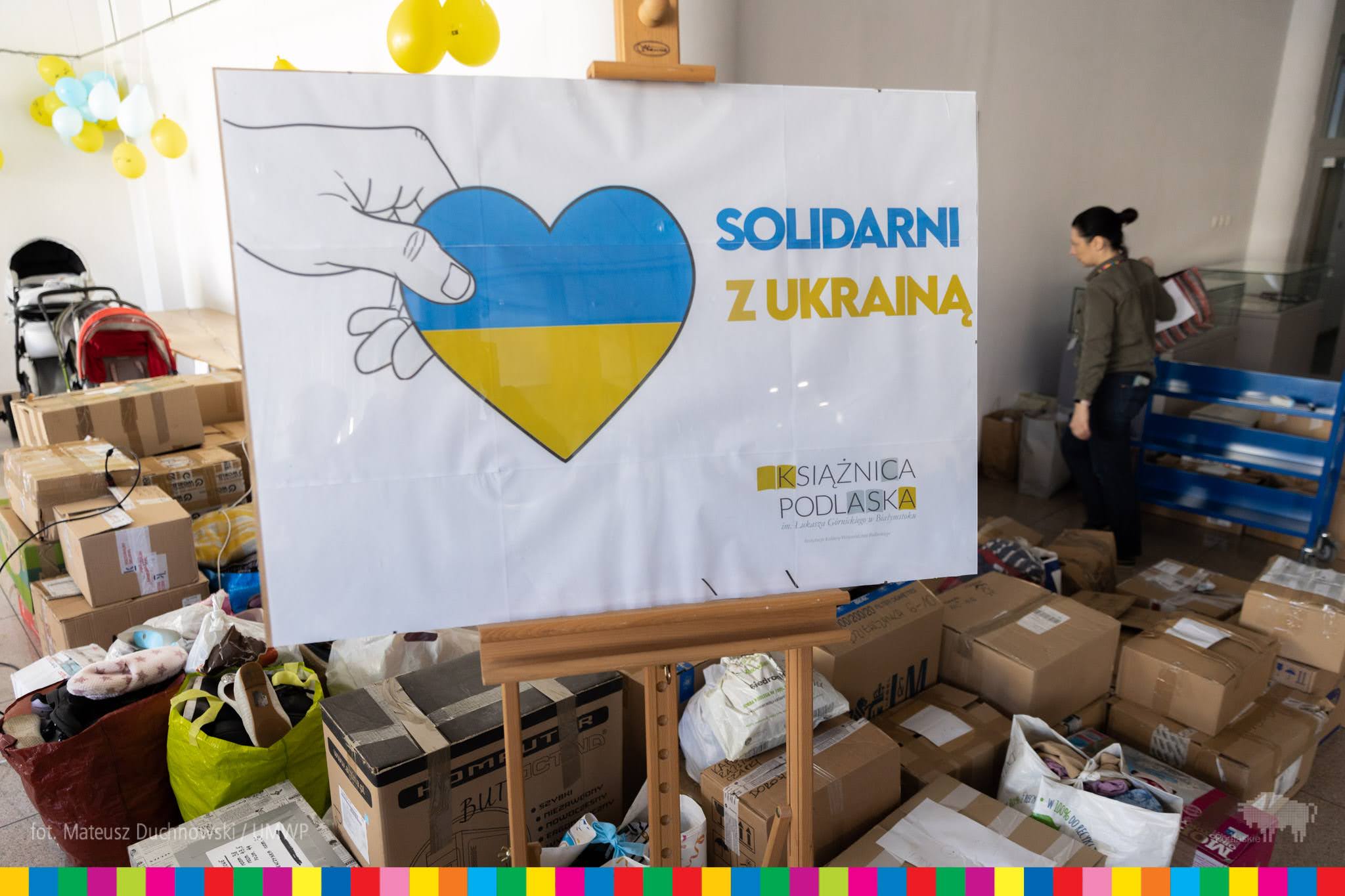 Na drewnianej sztaludze stoi plakat; na białym tle dłoń wystająca z lewej strony trzyma serce, które od góry do połowy figury jest niebieskie, a pod spodem żółte. Obok serca jest niebiesko-żółty napis : Solidarni z Ukrainą. Wokół sztalugi, znajdują się kartony z darami