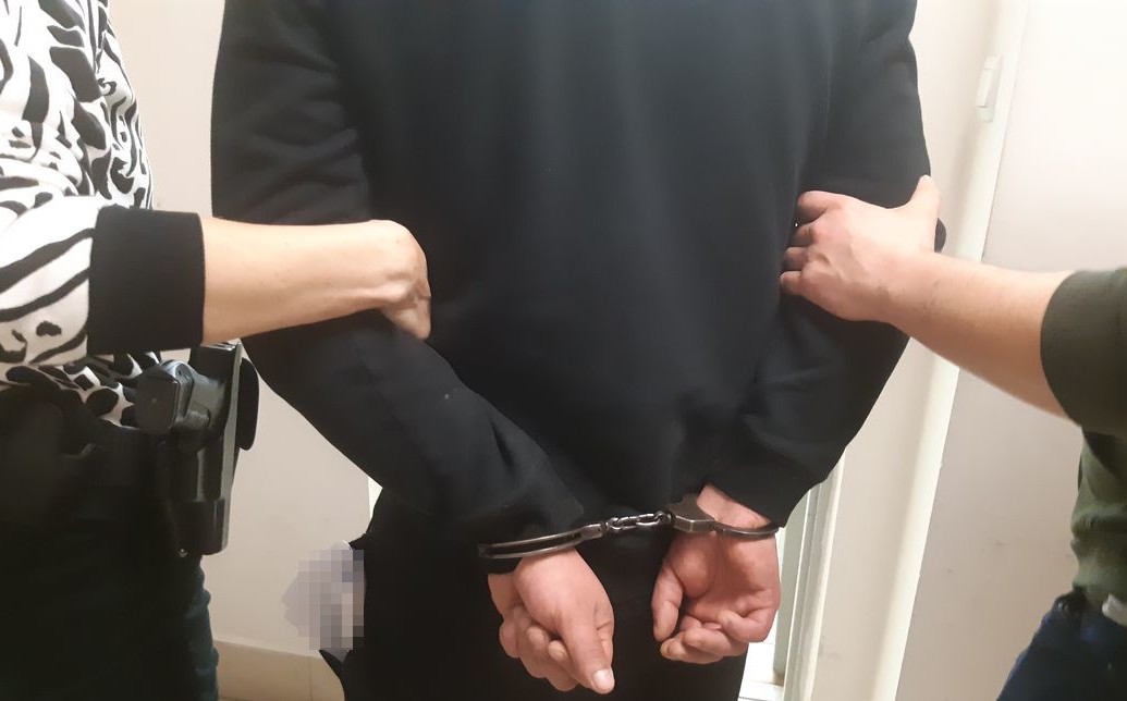 Mężczyzna w czarnej bluzie i spodniach ma z tyłu skute kajdankami ręce. Pod rękę z lewej i prawej strony trzymają go policjanci