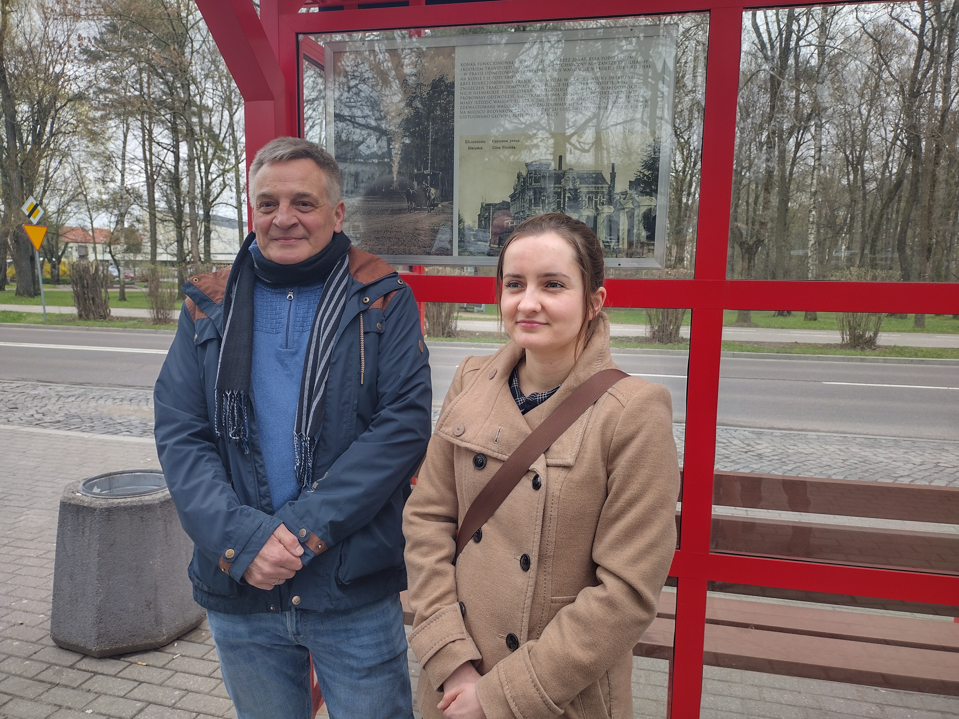 Na tle czerwonej wiaty autobusowej z przeszkleniami z lewej strony stoi mężczyzna, z prawej kobieta