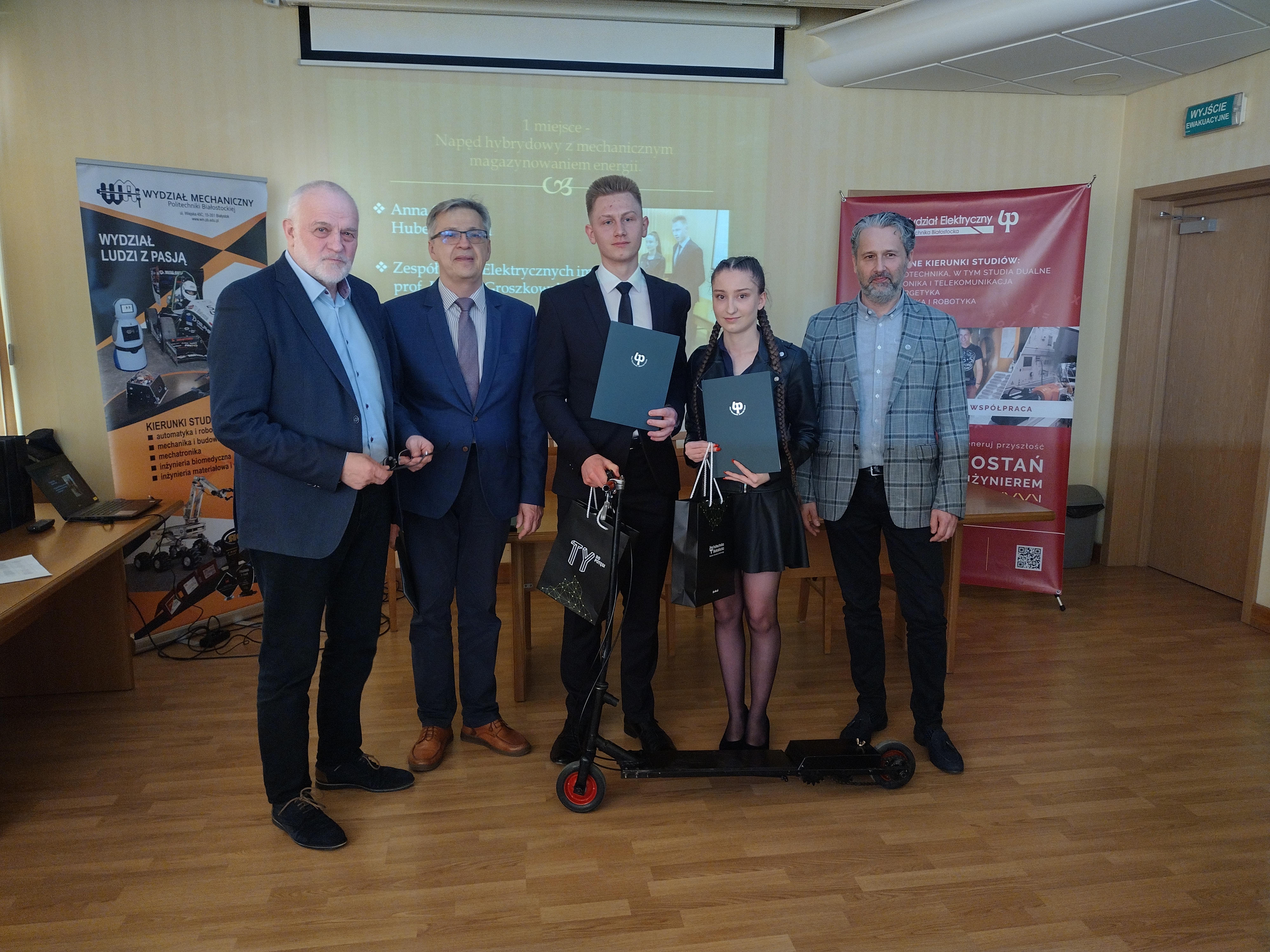Zdjęcie w sali, z lewej strony stoi dwóch mężczyzn, po środku chłopak z dziewczyną, a przed nimi hulajnoga, a z prawej stoi kolejny mężczyzna
