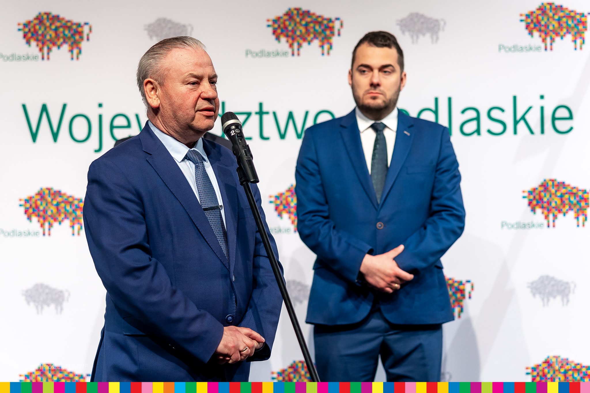 Mężczyzna w granatowym garniturze przemawia do mikrofonu. Stoi on na tle białej ścianki w pikselowe, kolorowe żubry z zielonym napisem: województwo podlaskie. Na tej ściance z prawej strony stoi drugi mężczyzna w garniturze w morskim kolorze