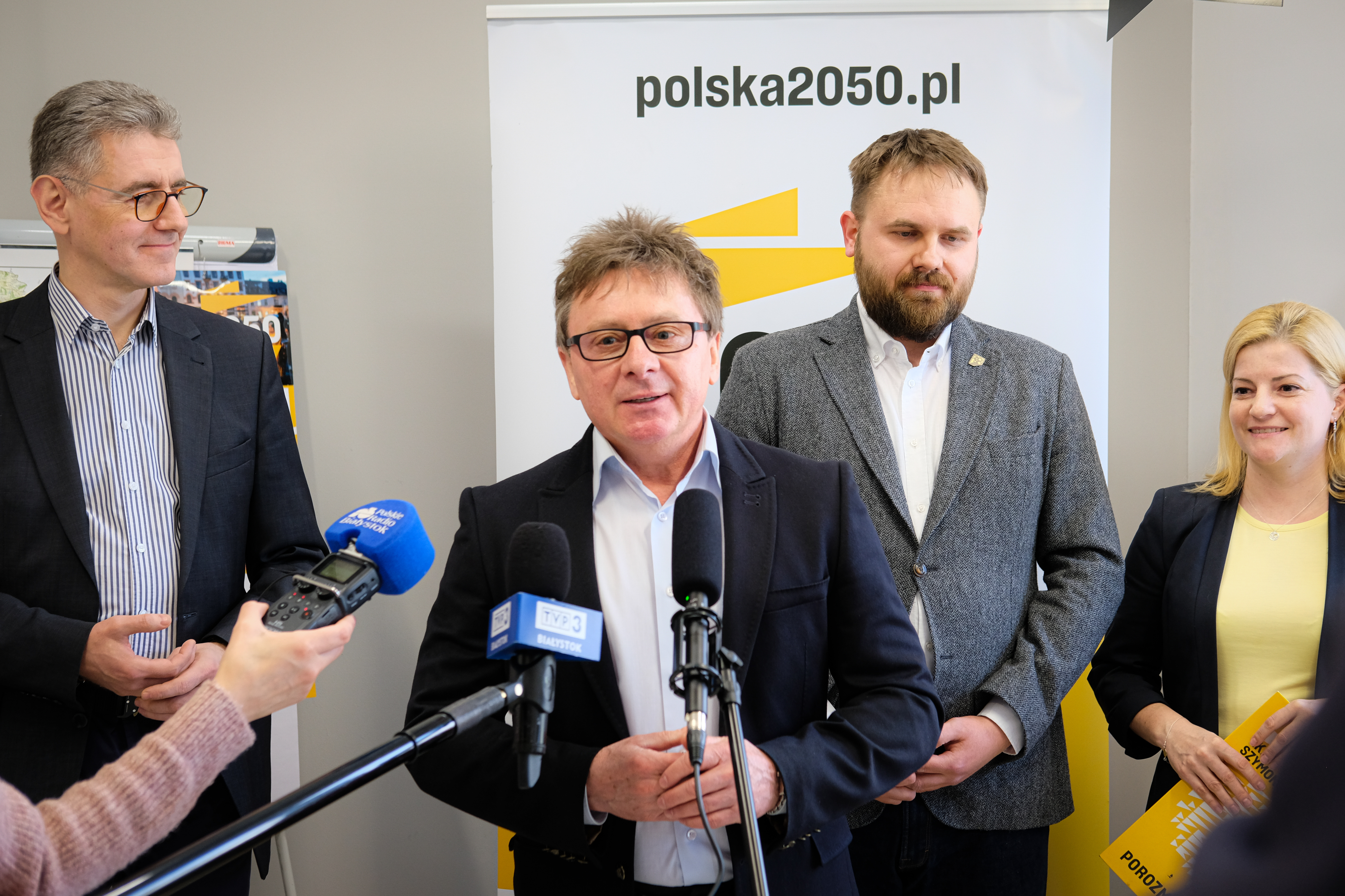 Niewysoki mężczyzna, w okularach mówi do mikrofonu, które przed nim trzymają dziennikarze. Za nim stoi jeszcze dwóch innych mężczyzn, z lewej strony i po środku, a z prawej stoi kobieta