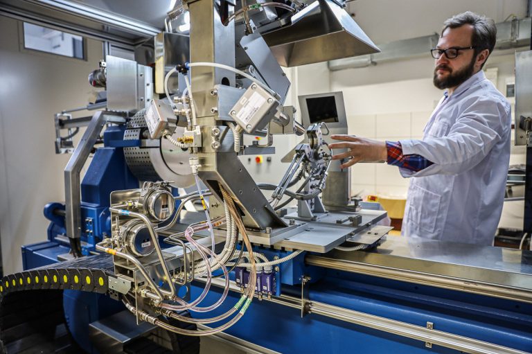 Maszyna w laboratorium, przy której stoi mężczyzna w białym fartuchu