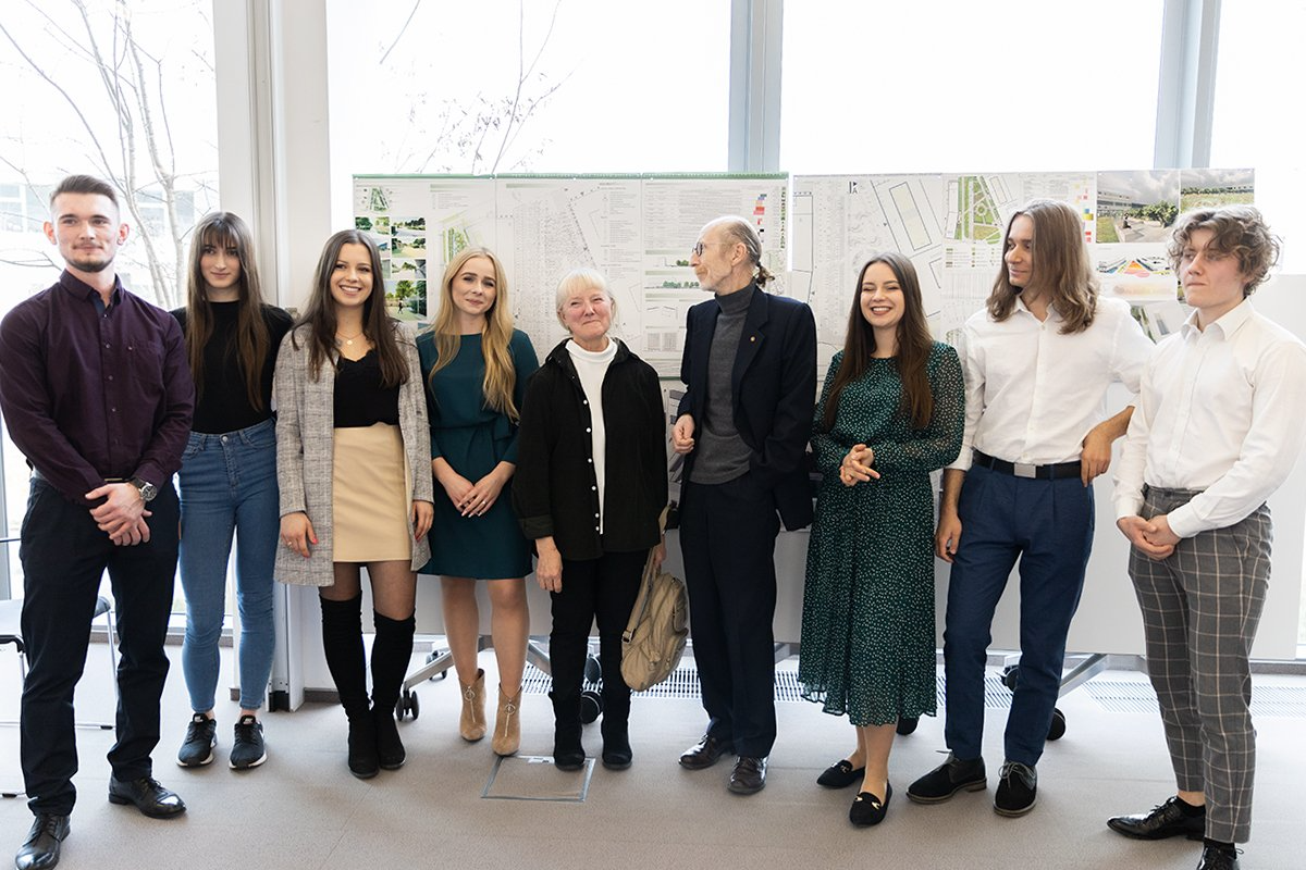 Na tle dużego okna stoi plansza z projektami, na której z kolei stoi grupa studentów