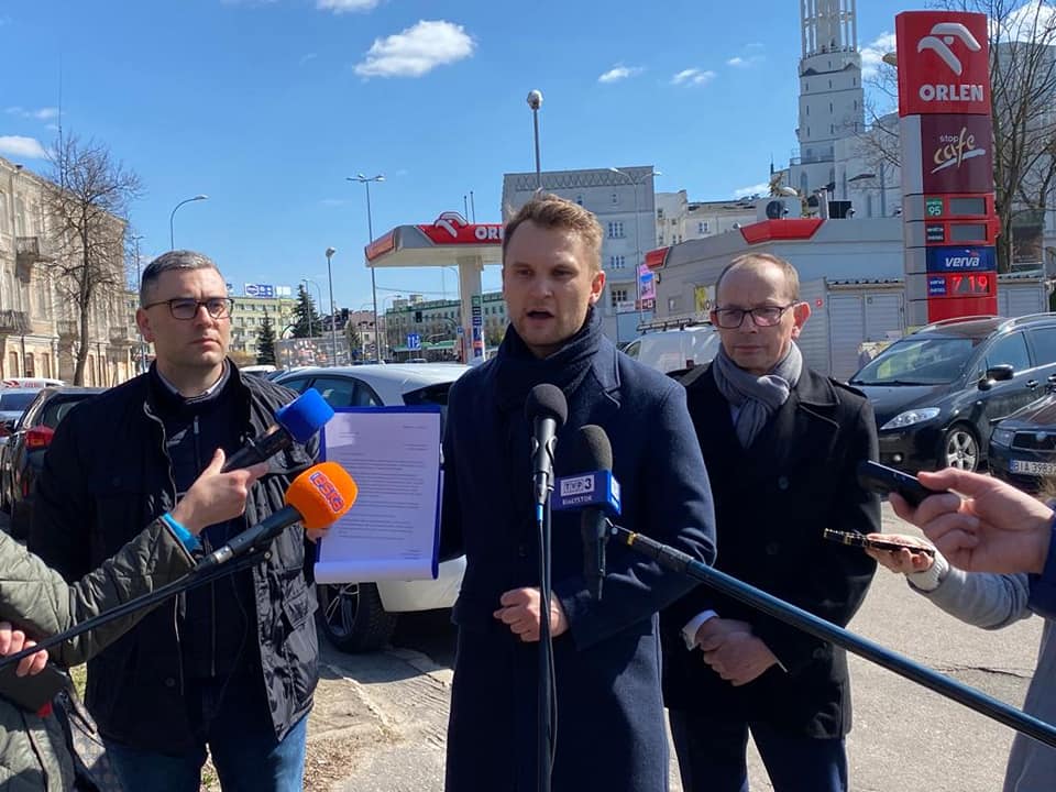 Słoneczny dzień. Trzech mężczyzn stoi na zewnątrz, stojący pośrodku jest wysunięty do przodu, bo mówi mikrofonów dziennikarzy. Ze swojej prawej strony trzyma białą kartkę z interpelacją. Za mężczyznami jest stacja paliw Orlen