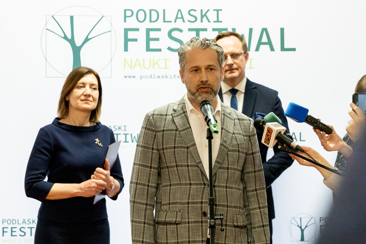 Mężczyzna w ciemno-beżowej marynarce w kratkę stoi przy mikrofonie, do którego mówi. Za nim z lewej strony stoi kobieta, z prawej mężczyzna