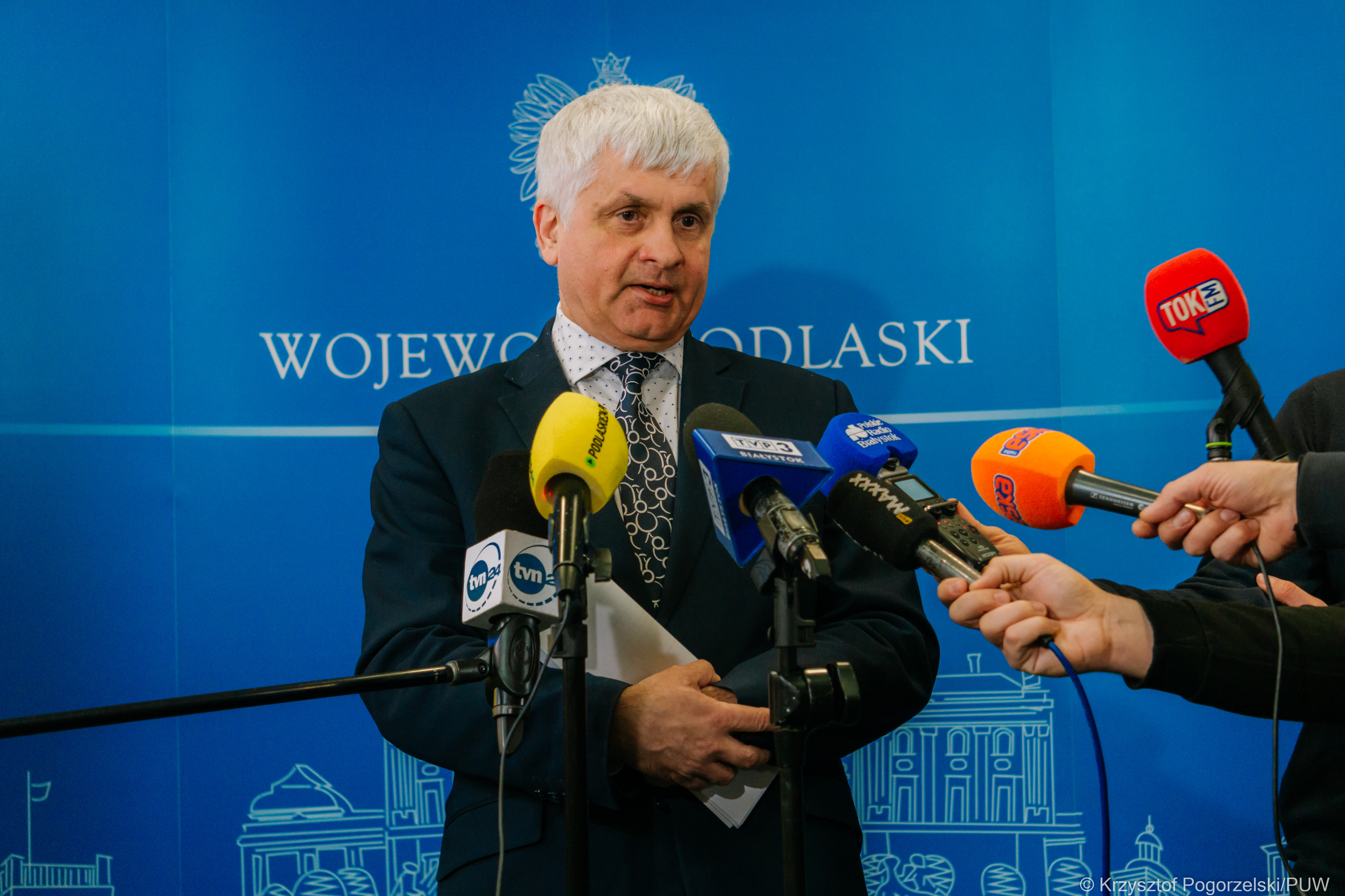 Na niebieskiej ściance z białym napisem: wojewoda podlaski, stoi siwy mężczyzna, w ciemnej marynarce i mówi do mikrofonów, które przed nim trzymają dziennikarze