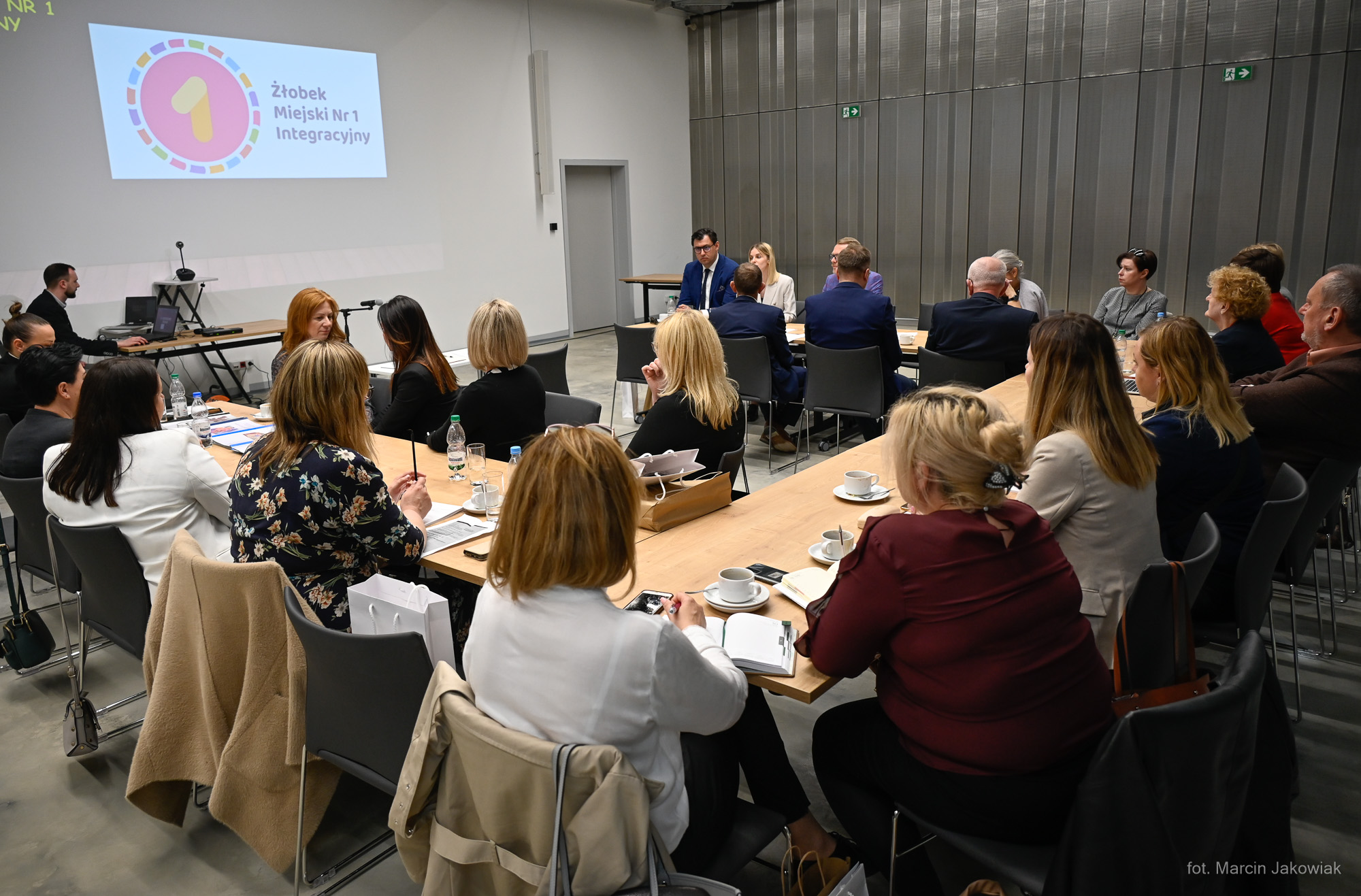 W sali konferencyjnej, przy podłużnych stołach, tyłem siedzą osoby;kobiety, mężczyźni. Patrzą na rzutnik, który znajduje się na ścianie, z lewej strony. Na ścianie jest wyświetlony napis: Przedszkole integracyjne nr 1 w Białymstoku