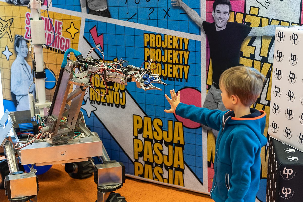 Z lewej strony stoi łazik marsjański, z prawej chłopiec w niebieskiej bluzie, który wyciąga do niego rękę