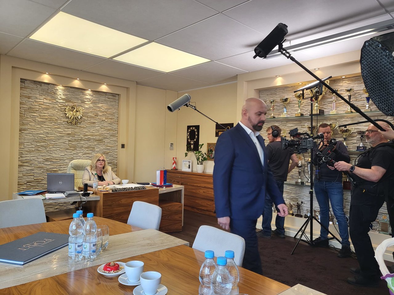 Scena z filmu, która dzieje się w gabinecie burmistrza. Za biurkiem, z lewej strony zdjęcia, siedzi kobieta. Blondynka w okularach leczniczych w czarnych oprawkach i ubrana na jasno. Na pierwszym planie widać stół konferencyjny. Przez salę idzie mężczyzna w granatowym garniturze. Z prawej strony widać ekipę filmowców, którzy mają kamery i mikrofony na wysięgnikach