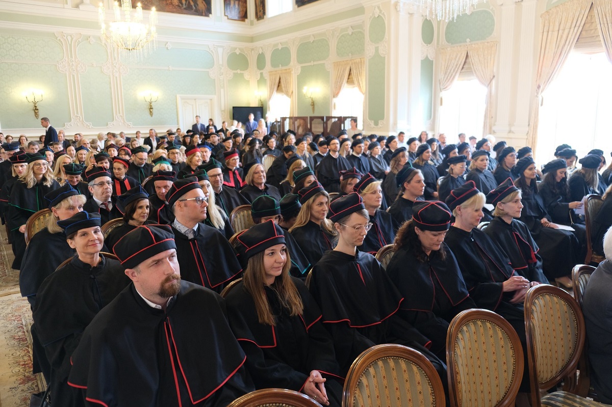 Kobiety i mężczyźni w czarnych togach siedzą w auli