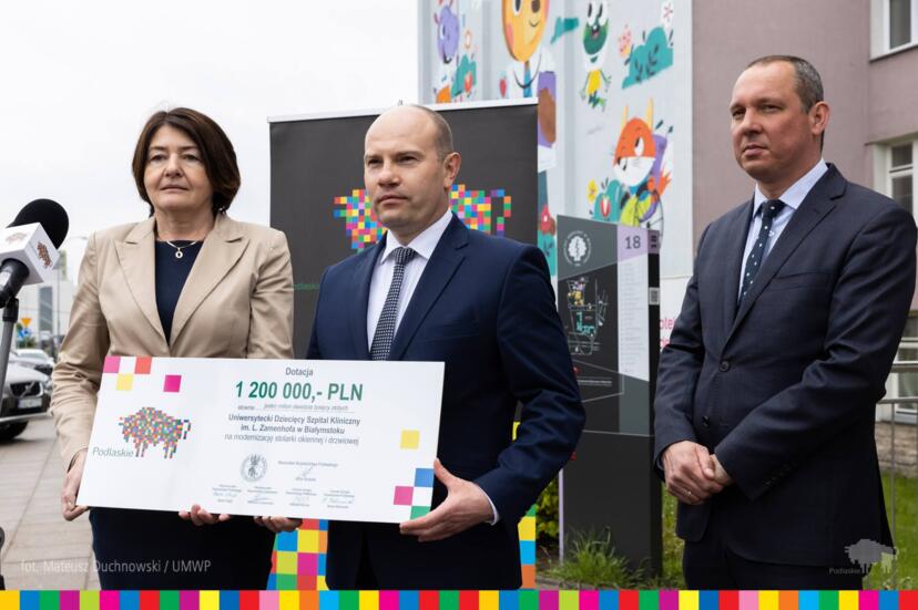 Trzy osoby przy szpitalu, jedna trzyma czek