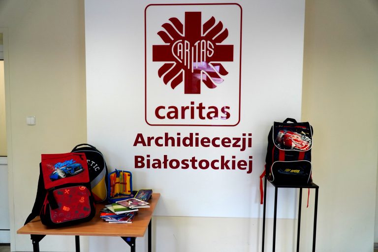 plakat Caritas Archidiecezji Białostockiej a po dwóch stronach plecaki szkolne i zeszyty