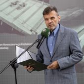 Mężczyzna elegancko ubrany przemawia do mikrofonu, trzyma otwartą teczkę