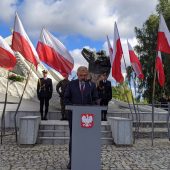 Mężczyzna w garniturze stoi przed mównicą, za nim jest pomnik, flagi Polski i żołnierze