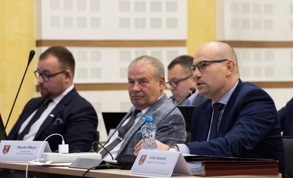 po prawej widoczny jest mężczyzna w garniturze i okularach mowiący do mikrofonu, pośrodku mężczyzna w jasnym garniturze, po lewej mężczyzna w ciemnym garniturze pracujący na komputerze