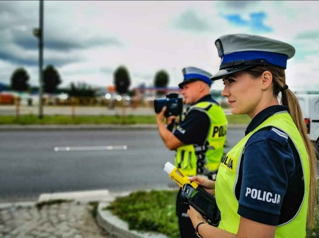 Policjant drogówki trzyma przy twarzy miernik laserowy, obok stoi policjantka trzymająca w ręku alkomat.