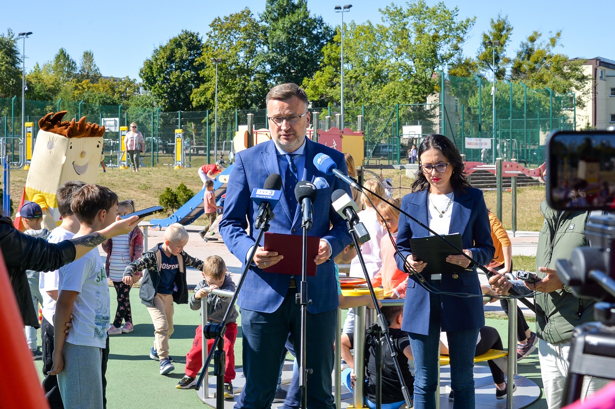 Konferencja prasowa na placu zabaw. Do mikrofonów dziennikarzy przemawia mężczyzna w garniturze.