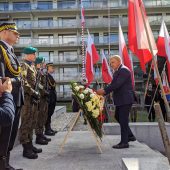 Mężczyzna w garniturze dotyka wieńca z kwiatami, po lewej stoją żołnierze na baczność, po prawej widać flagi Polski