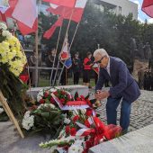 Starszy mężczyzna składa kwiaty obok innych wieńców, nad nim widać flagi Polski