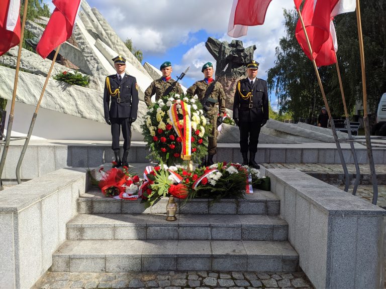 Czterech umundurowanych żołnierzy stoi przy pomniku, przed nimi leżą wieńce z kwiatami