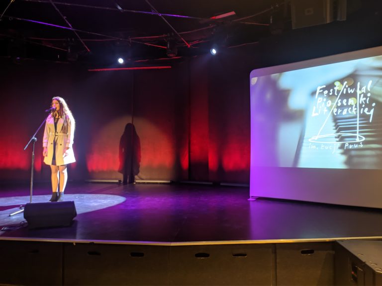 Kobieta stoi na scenie przed mikrofonem, obok na ekranie wyśwuetla się plakat festiwalu