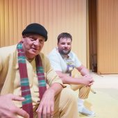 Dwóch mężczyzn siedzi na scenie, jeden unosi rękę jakby coś mówił