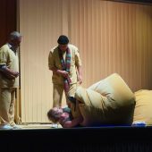 Na scenie teatralnej dwóch mężczyzn stoi nad jednym leżżacym na scenie