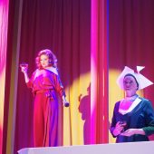 Jedna kobieta w eleganckiej sukni stoi z kieliszkiem wina i mikrofonem w ręku, druga kobieta ubrana jak zakonnica stoi ze skrzyżowanymi rękoma