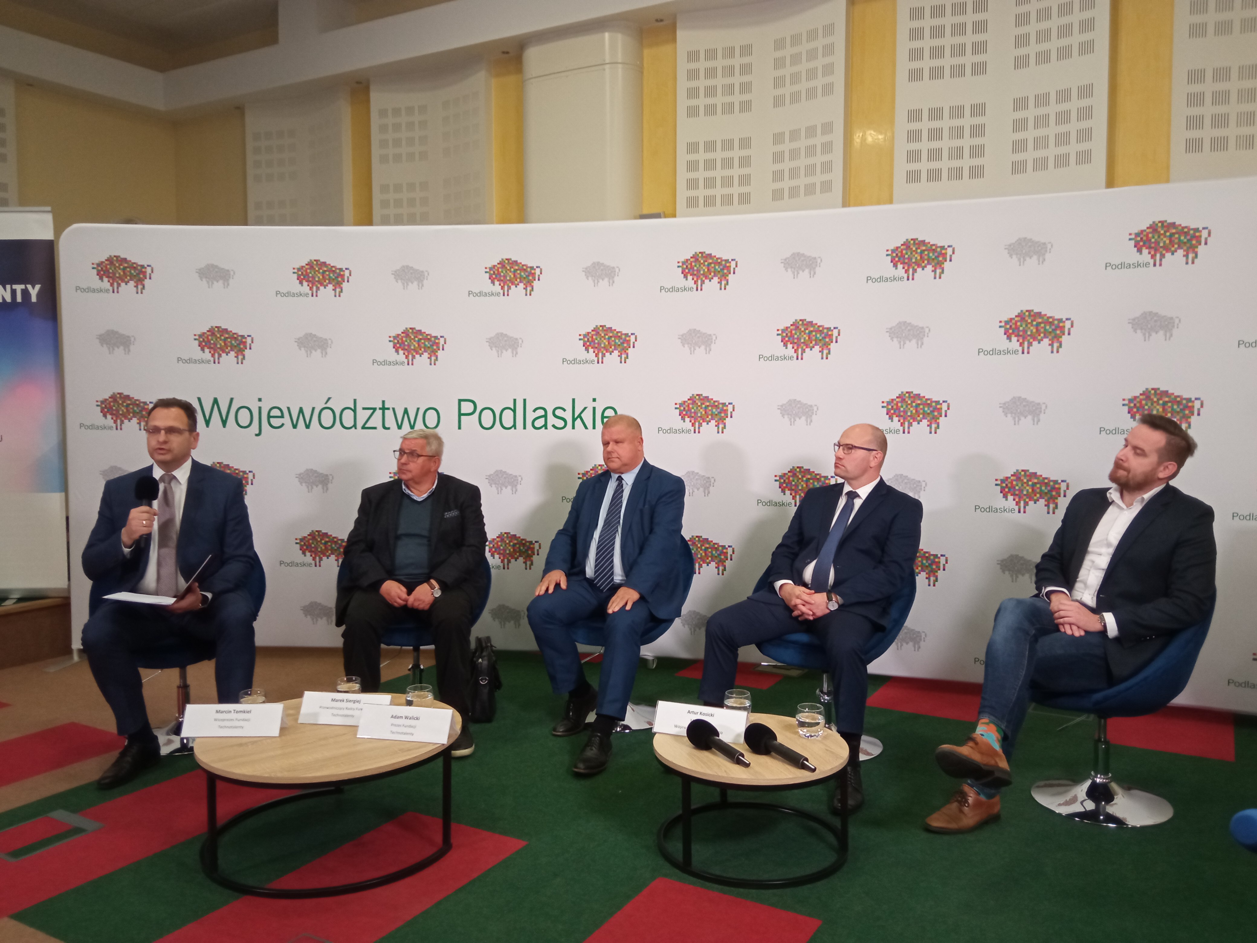 Pięciu mężczyzn sedzi na konferencji prasowej przy stolikach z mikrofonami
