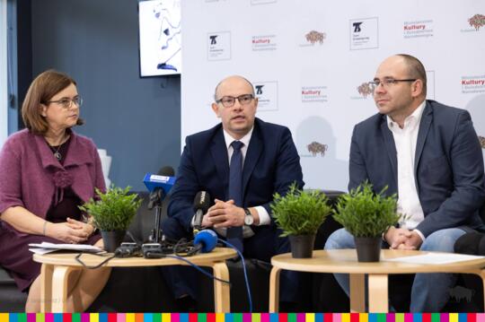 Dwóch mężczyzn i kobieta siedzą przy niskich stolikach z kwiatkami i mikrofonami.