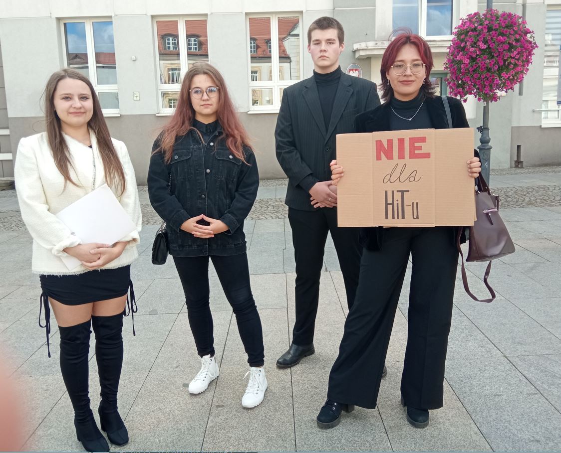 Trzy młode kobiety z kartonowym napisem i jeden mężczyzna stoją przed budynkiem kuratorium na Rynku Kościuszki