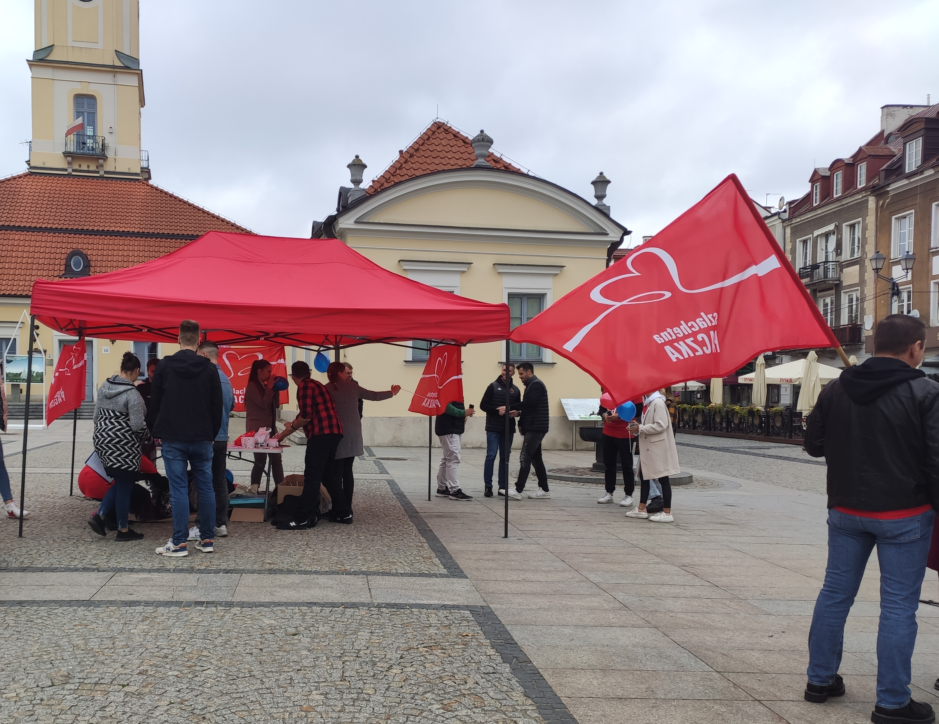 Przed ratuszem stoją ludzie pod czerwonym namiotem, wisi flaga z napisem Szlachetna Paczka