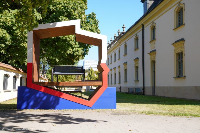 Przestrzenna instalacja w kształce mapy Polski, pomalowana na biało-czerwono stoi na platformie granatowej, w środku stoi ławeczka