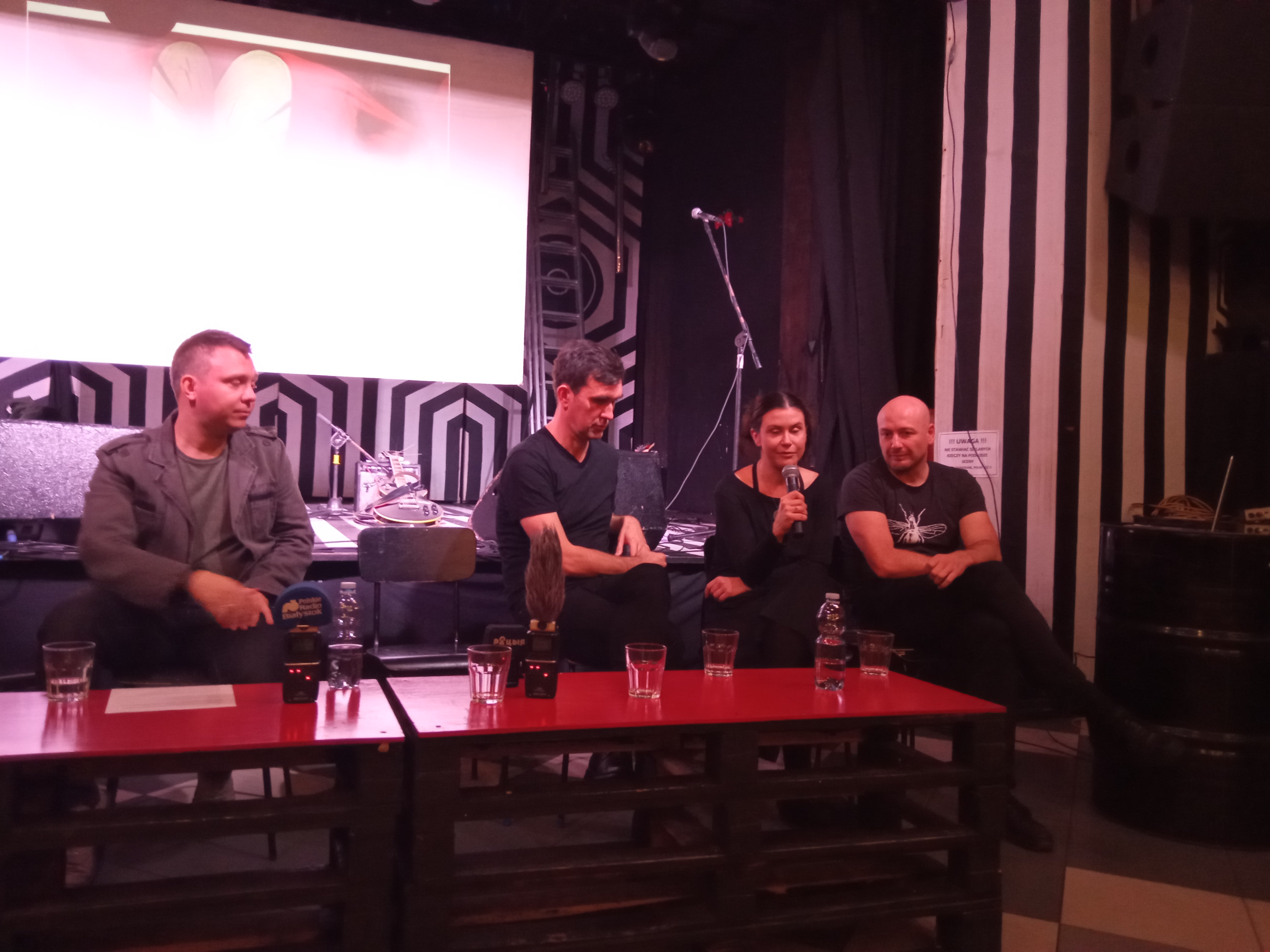 Kobieta i trzechch mężczyzn siedzi przy stoliku. Kobieta mówi do mikrofonu