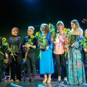 Grupa ludzi stoi obok siebie na scenie z dyplomami i słonecznikami