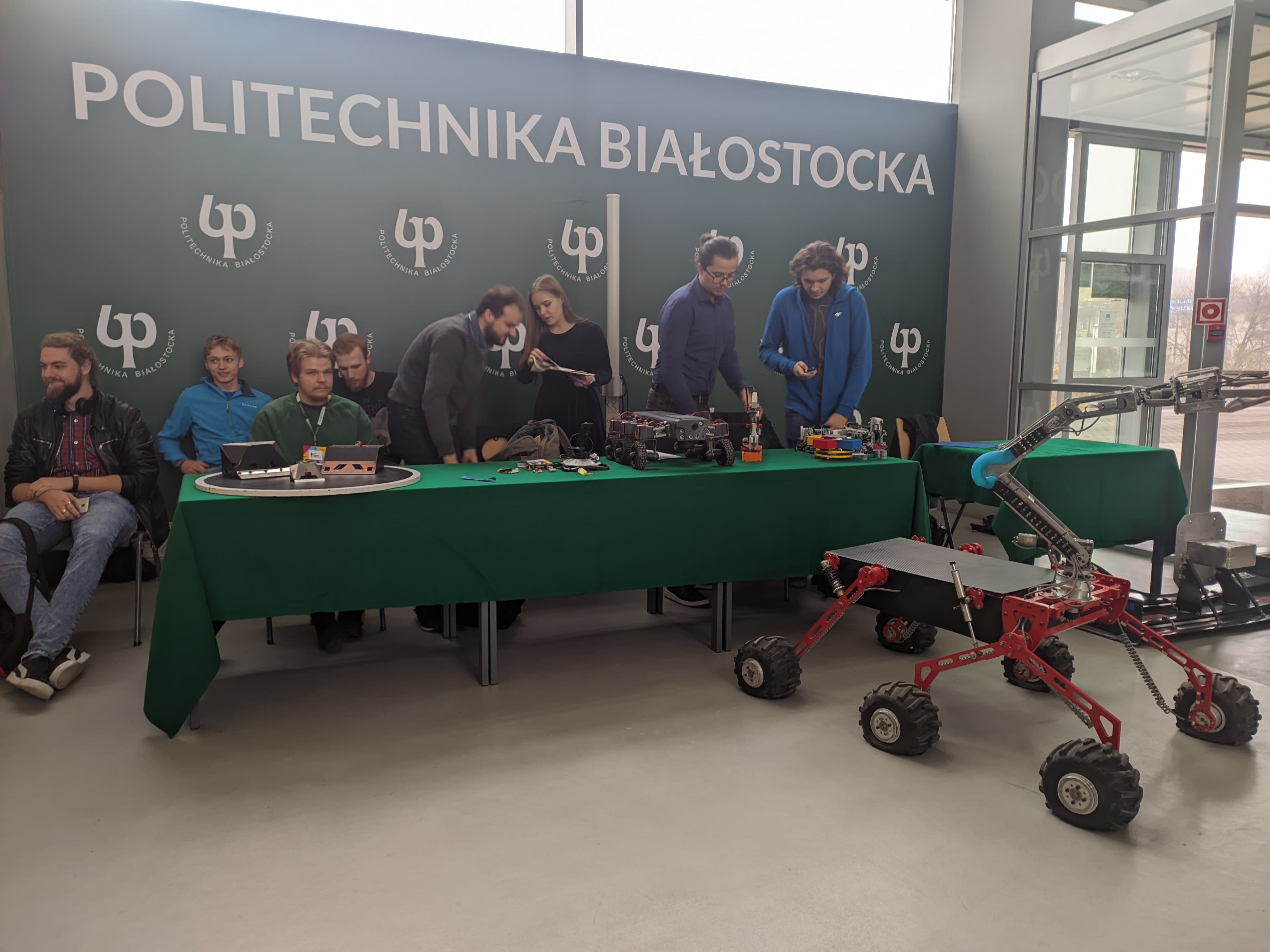 Młodzi ludzie stoją i siedzą za stołem, przed nimi jest łazik marsjański