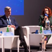 Kobieta siedząc na scenie w fotelu trzyma mikrofon przy ustach, obok niej siedzi mężczyzna