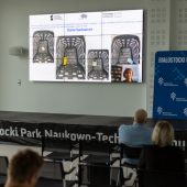 Mężczyzna prezentuje na dużym ekranie projekt, na krzesłach siedzą słuchacze