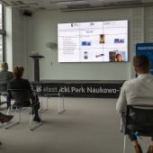 Mężczyzna prezentuje na dużym ekranie projekt, na krzesłach siedzą słuchacze