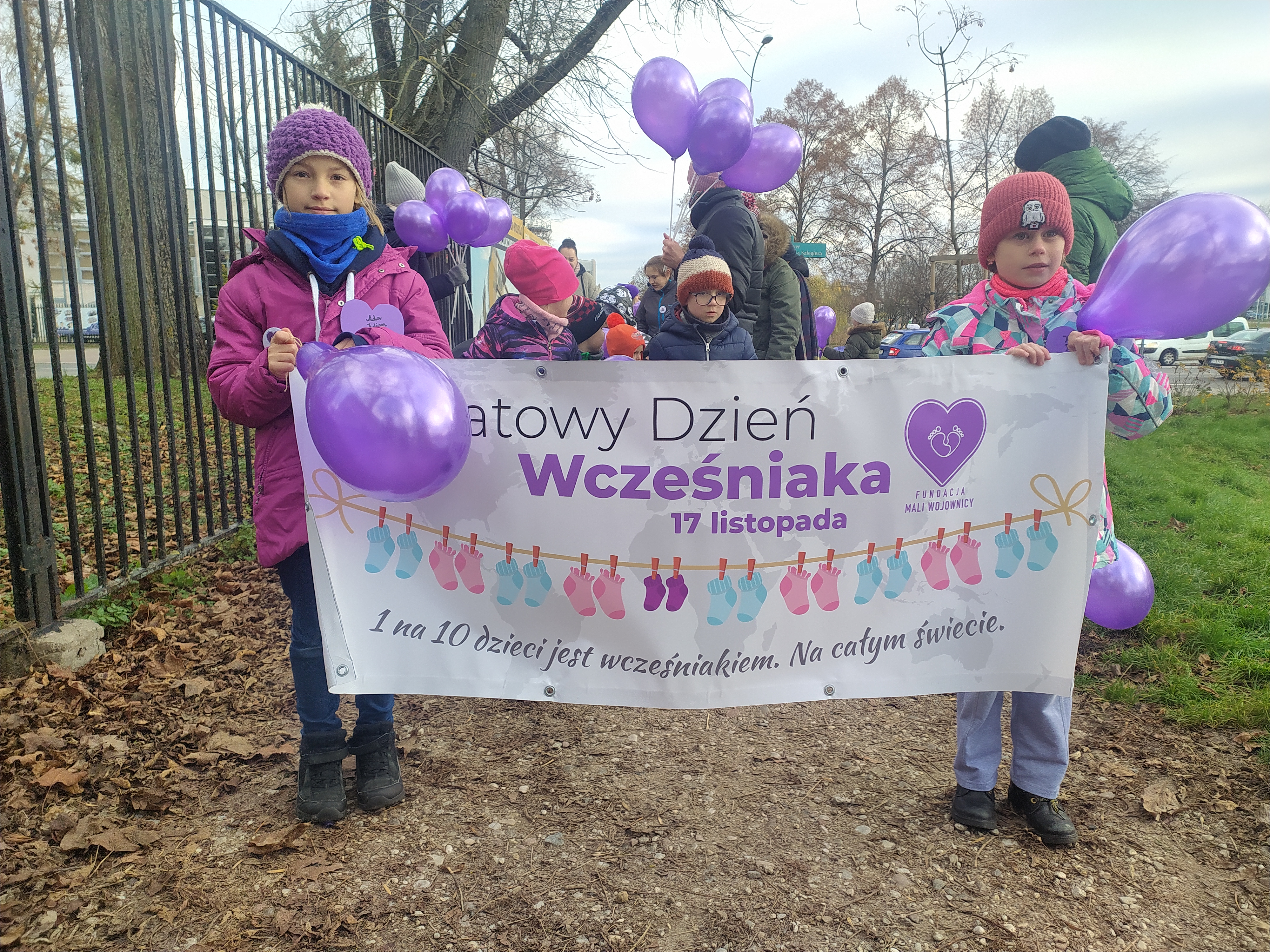 Dziewczynki trzymają transparent z napisem Światowy Dzień Wcześniaka, za nimi dzieci trzymają fioletowe balony