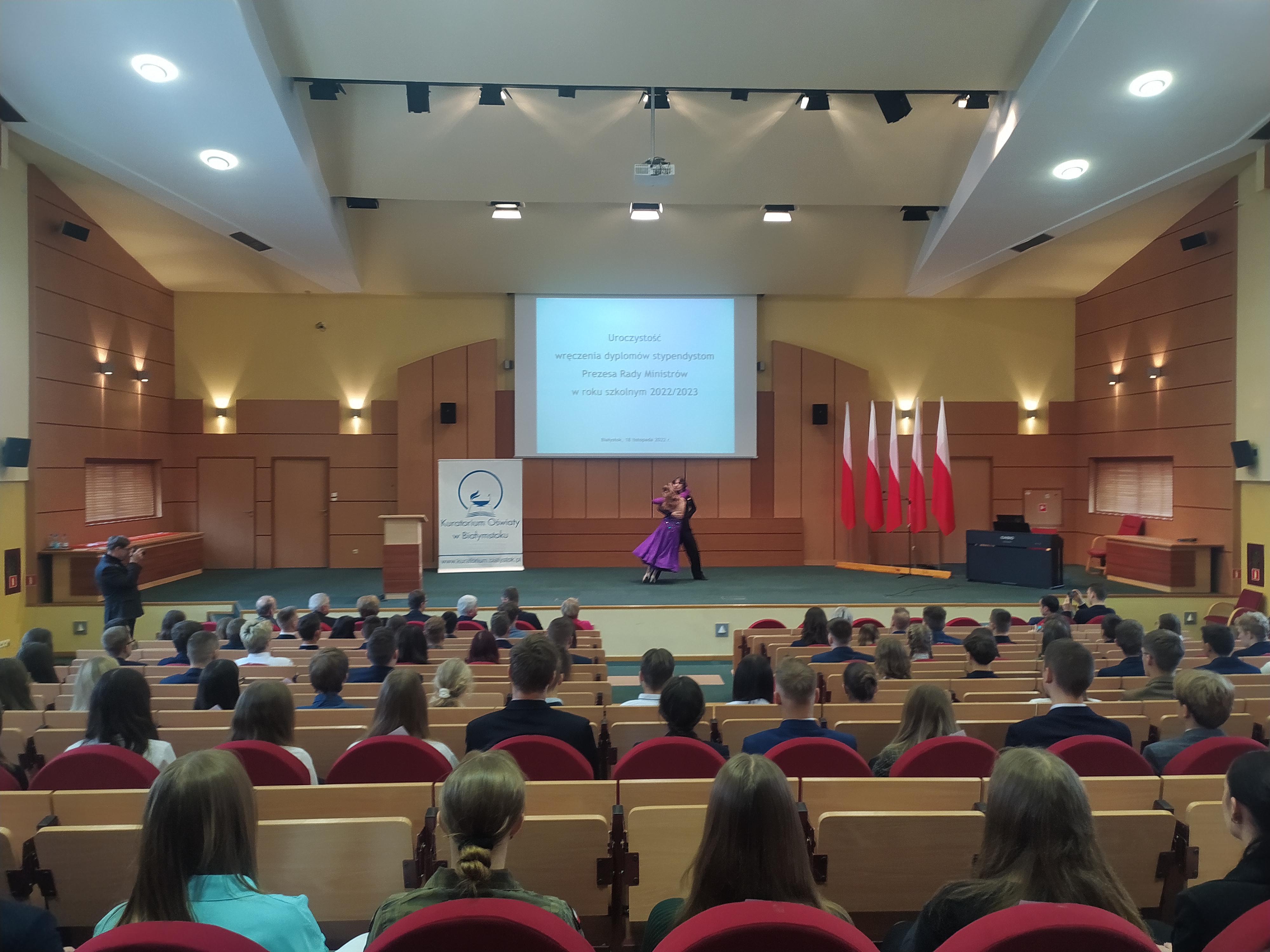 Aula, na której widzimy siedzącycy tyłem do nas młodych ludzi, na scenie jest tańcząca para.