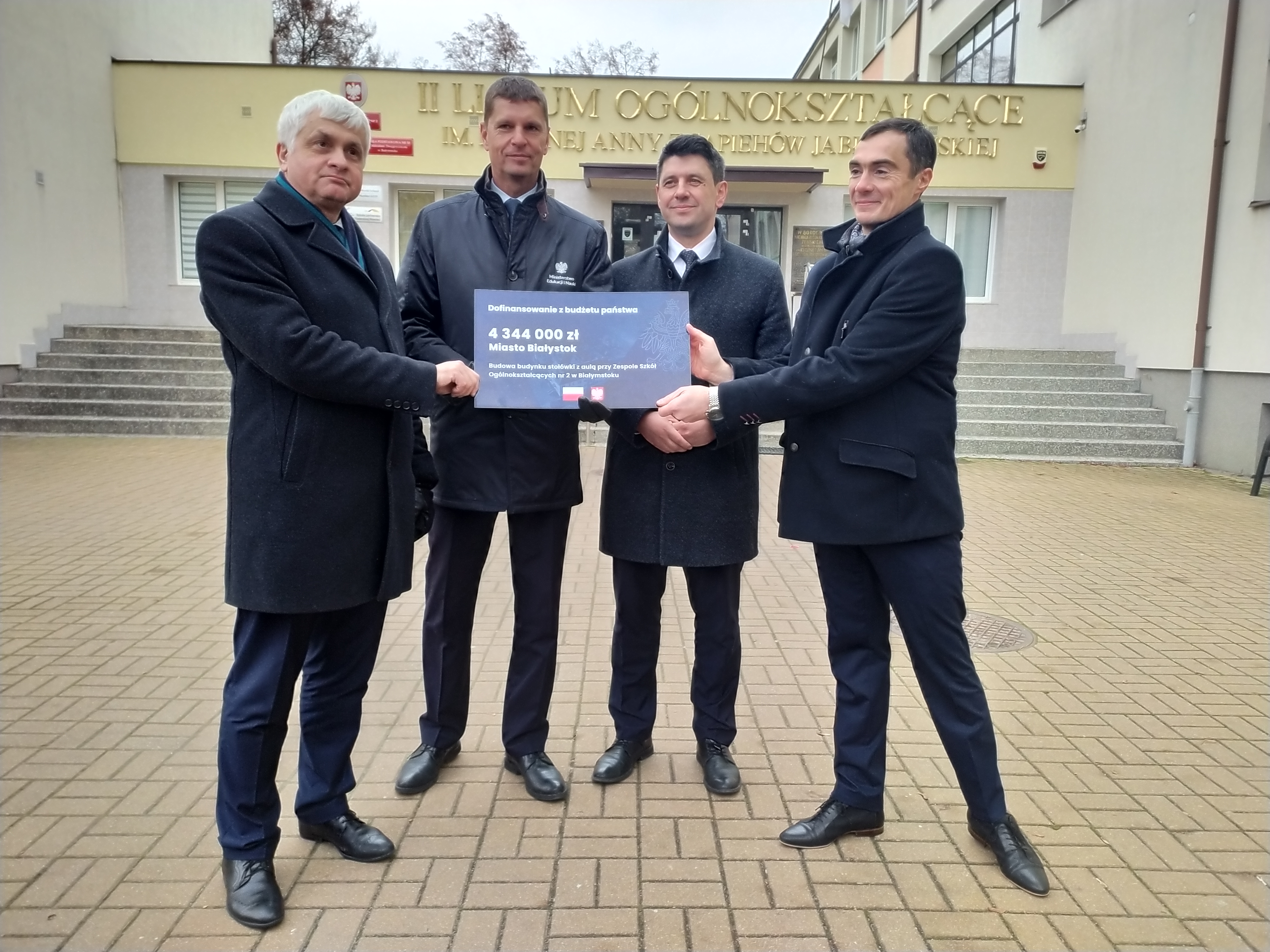 Czterech mężczyzn trzyma symboliczny czek przed budynkiem szkoły