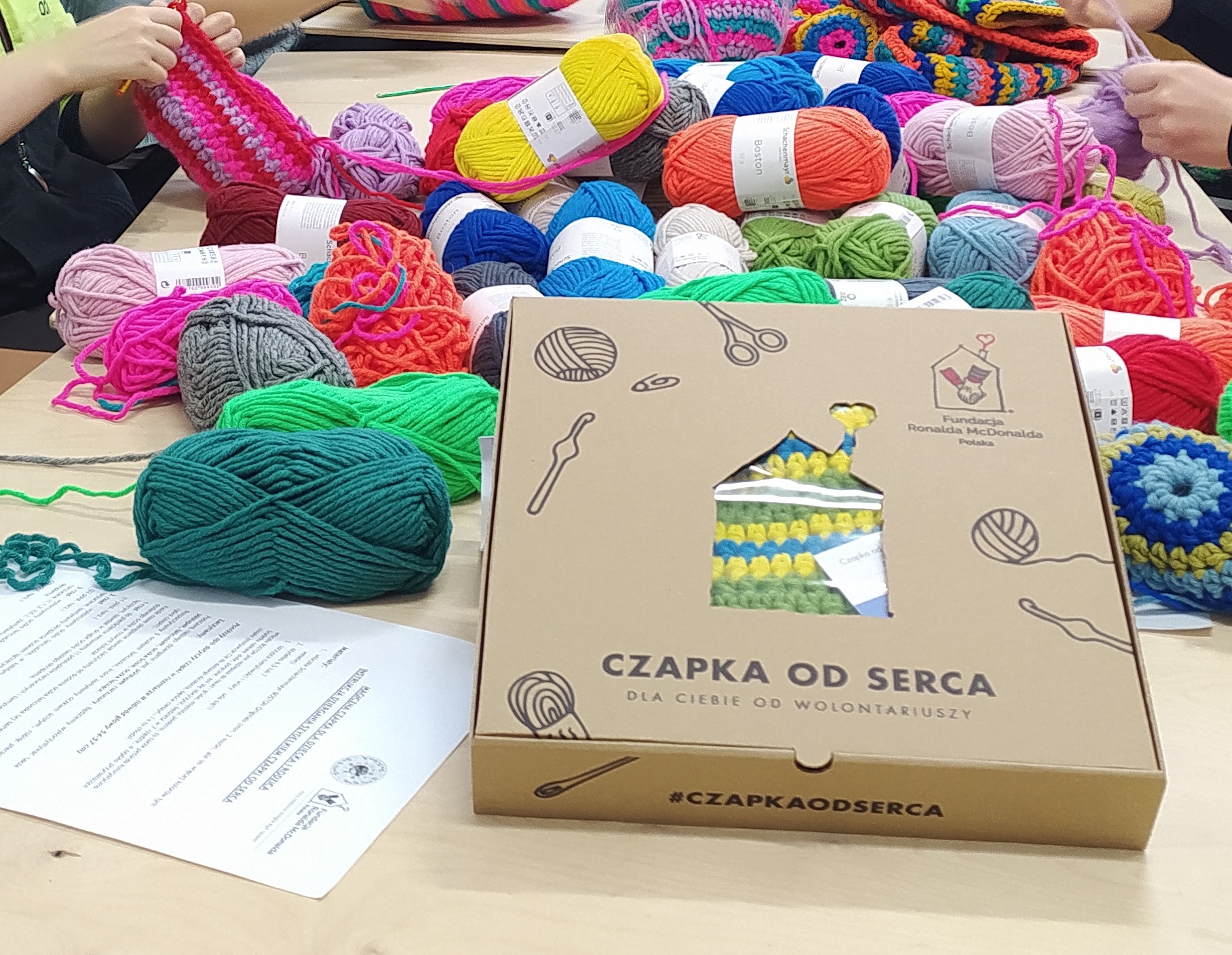 Pudełko z napisem "Czapki od serca" a za nim na stole leżą kolorowe wlóczki