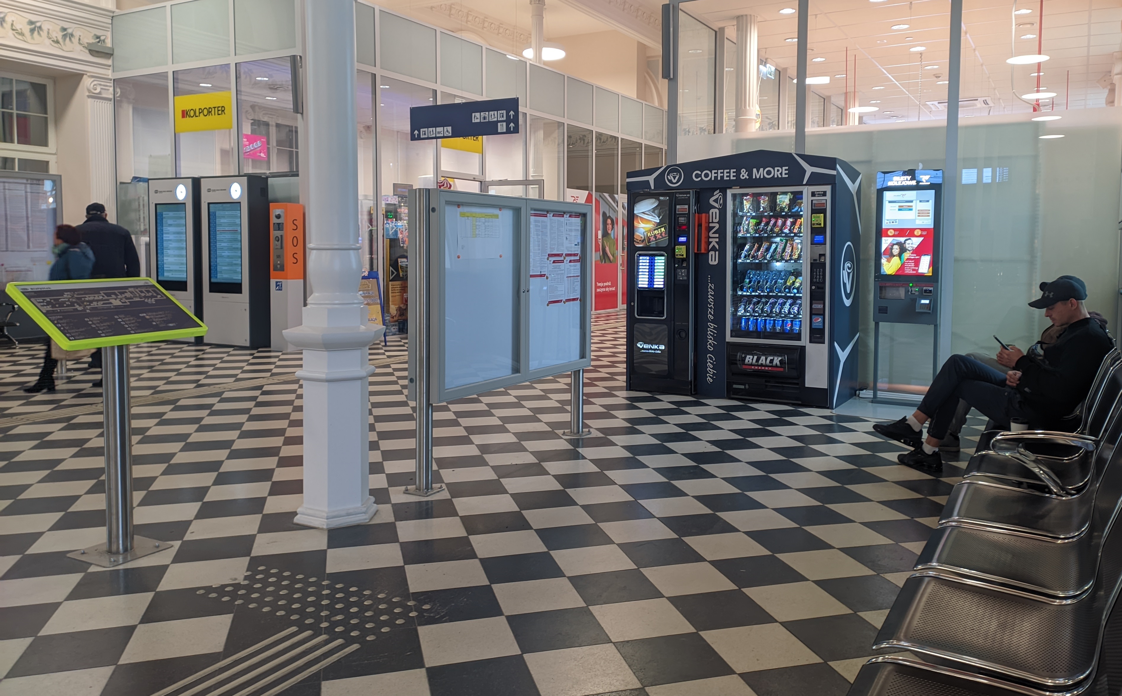 Wnętrza dworca, na którym widać tablicę z rozkładem jazdy, automat z przekąskami, videomat i siedzących ludzi